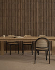 A minimalist dining room featuring the Wooden Stories Oli Dining Table, six black-framed chairs with woven seats, a closed book, a cup, and a dark wood-paneled wall in the background.