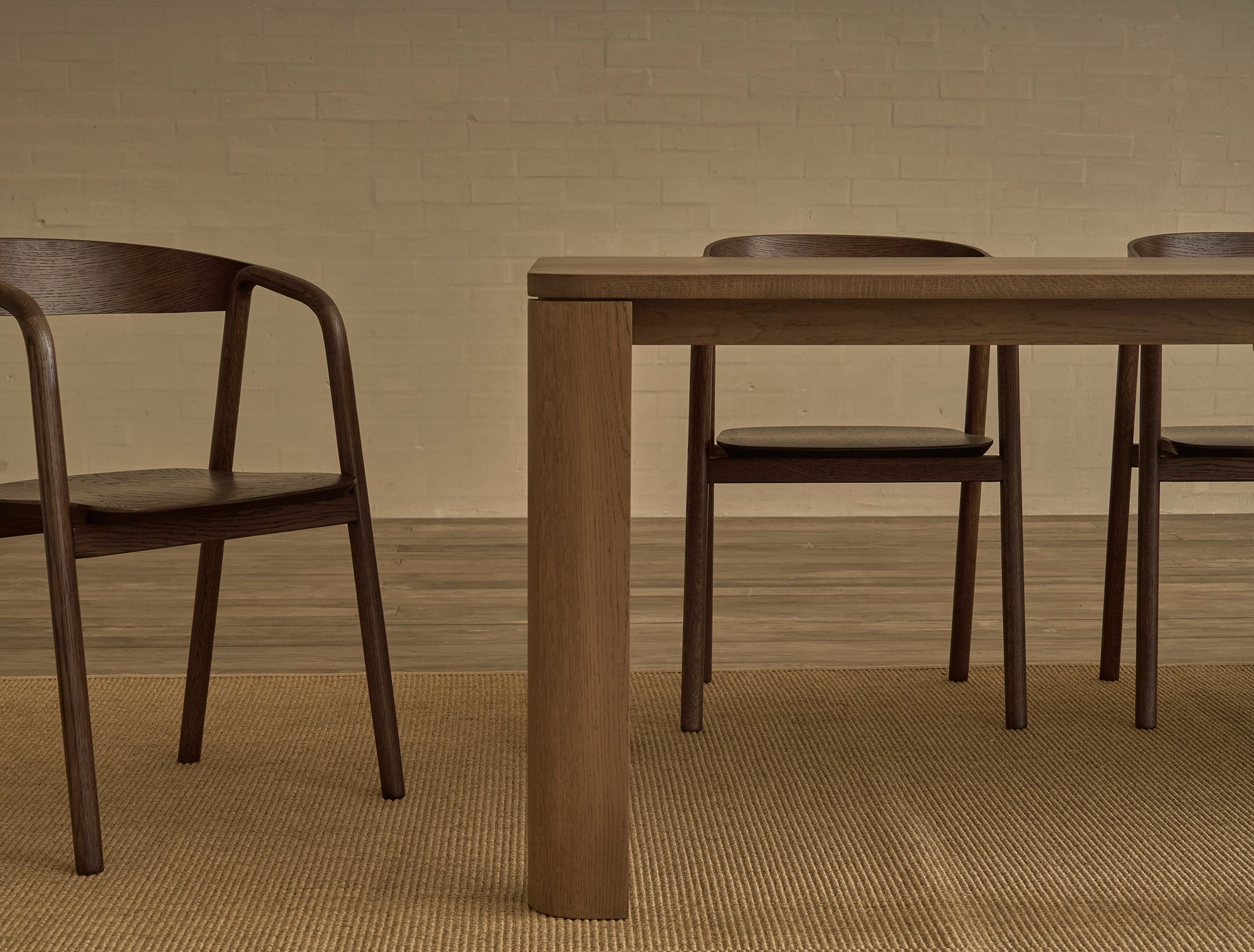 Two wooden chairs and the Oli Dining Table by Wooden Stories sit on a beige rug against a light brick wall, creating a minimalistic room that reflects the timeless elegance of Danish design.
