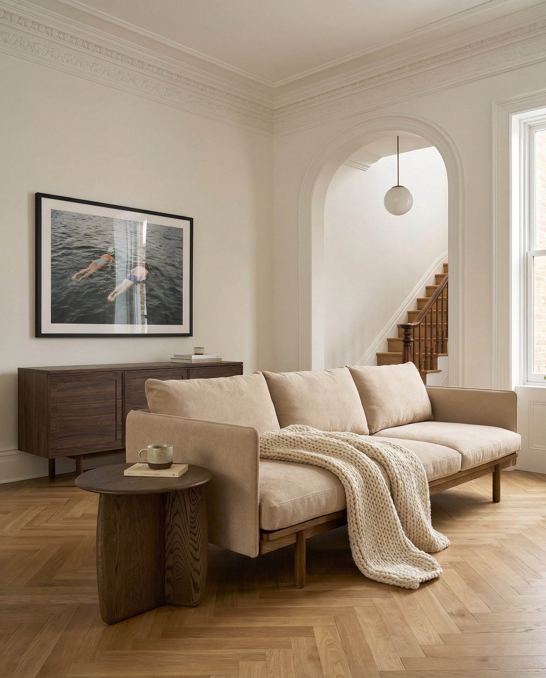 A neutral living room featuring the Wooden Stories Pensive Sofa | Fabric, chunky knit blanket, wooden side table with mug, framed artwork, wooden console, large window, and arched doorway to a staircase.
