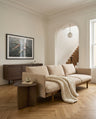 A neutral living room featuring the Wooden Stories Pensive Sofa | Fabric, chunky knit blanket, wooden side table with mug, framed artwork, wooden console, large window, and arched doorway to a staircase.