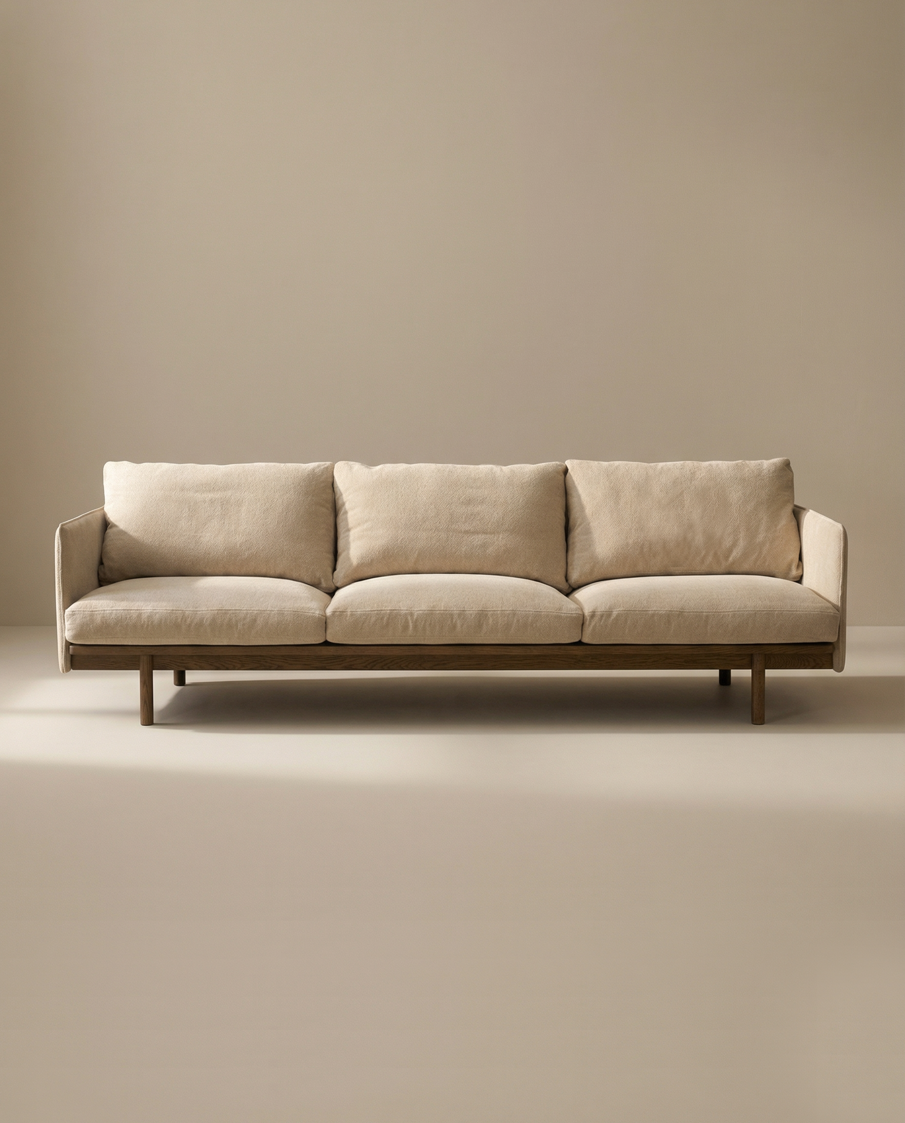 A minimalist beige three-seater Pensive Sofa | Fabric by Wooden Stories, with plush cushions and a wooden base inspired by mid-century design, is set against a plain, light-toned background.