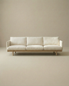 The Pensive Sofa | Fabric by Wooden Stories is a modern, minimalist Danish cream sofa with three seat cushions and wooden legs, shown against a plain beige background.