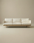 The Pensive Sofa | Fabric by Wooden Stories is a modern, minimalist Danish cream sofa with three seat cushions and wooden legs, shown against a plain beige background.