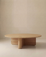 The Wooden Stories Pivot Coffee Table | Round features a minimalist Danish design with thick, geometric legs and a natural solid wood finish. Its modern, sculptural look stands out against a smooth, beige backdrop.