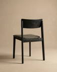 The Poise Dining Chair by Wooden Stories features a minimalist black oak frame and textured fabric seat, set against a neutral beige background and angled slightly away from the viewer.