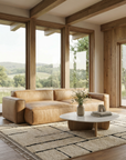 A modern living room with large windows, a tan leather sofa, a round coffee table, the SOBU Pasha Moroccan Rug, wooden floors, and scenic views of green hills outside.