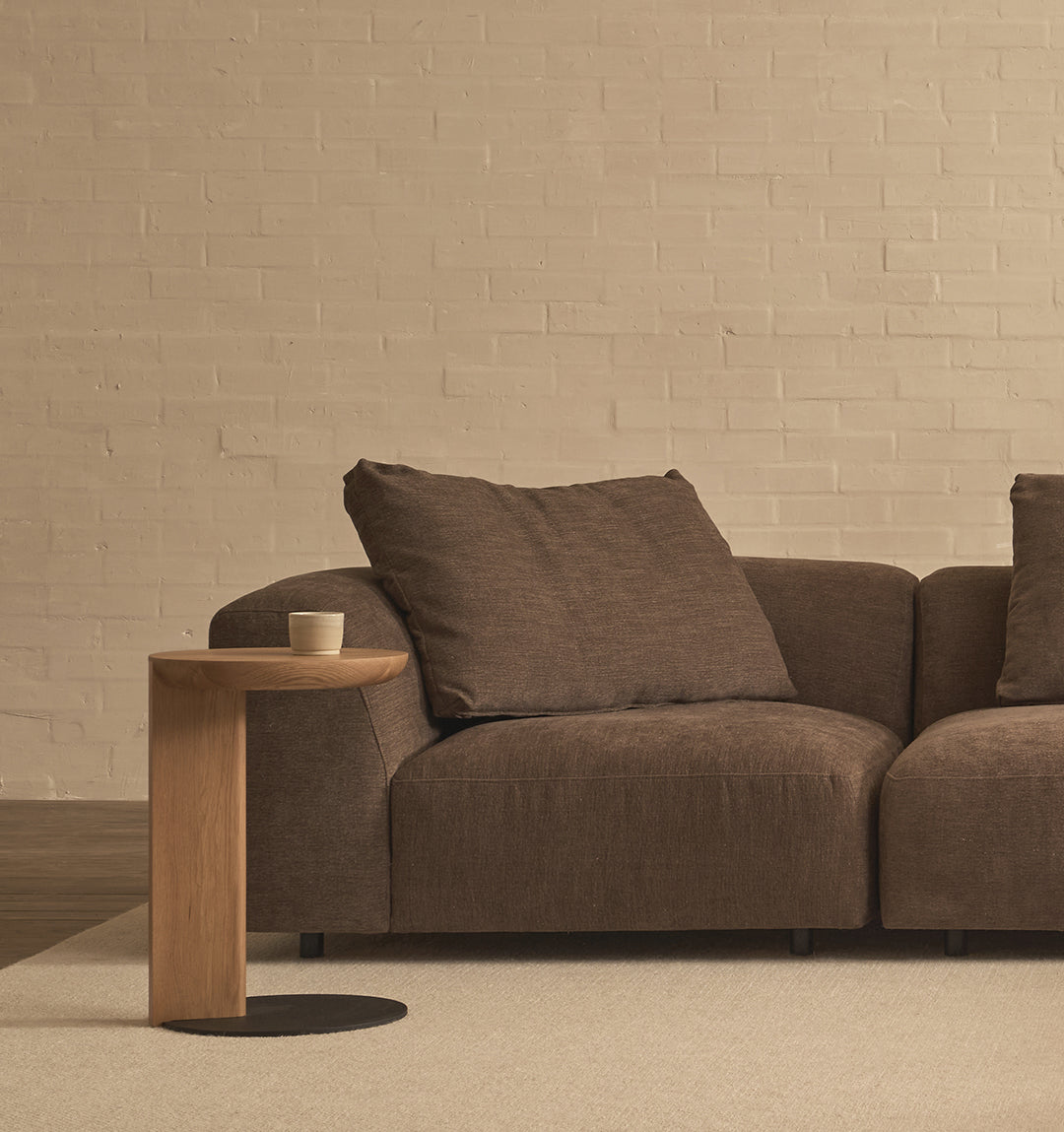 A modern living room with a brown fabric sofa, a beige carpet, a light brick wall from the Danish Collection, and a solid oak Wooden Stories Guild Side Table holding a cup.