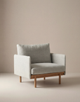 The Pensive Lounge Chair | Fabric by Wooden Stories features light grey upholstery, a wooden base, and clean mid-century lines, creating a simple, contemporary look in a minimalist room with neutral beige walls and floor.