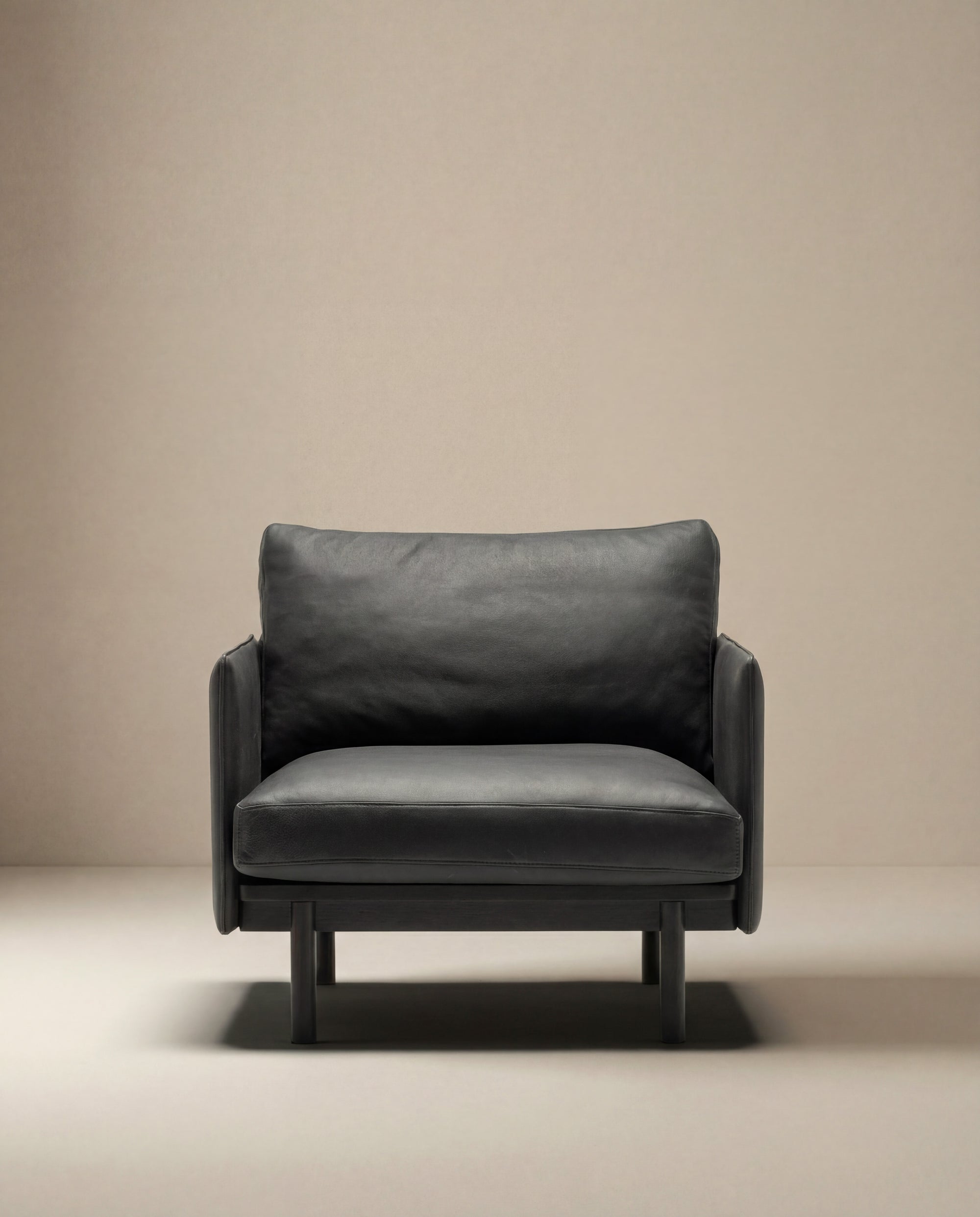 The Pensive Lounge Chair | Leather by Wooden Stories, a modern black leather seat with minimalist mid-century Danish design, sits on a light floor against a plain beige wall.