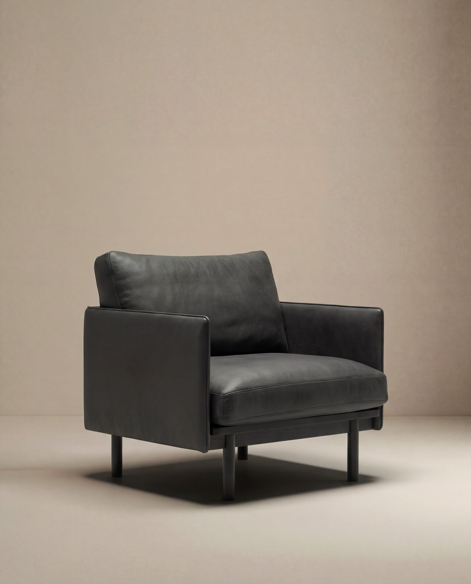 The Pensive Lounge Chair | Leather by Wooden Stories features a modern, black leather design with wide armrests and a boxy silhouette, inspired by mid-century Danish style. It sits on a light beige floor with a matching beige background.