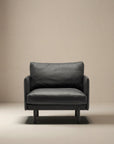 The Pensive Lounge Chair | Leather by Wooden Stories, a modern black leather seat with minimalist mid-century Danish design, sits on a light floor against a plain beige wall.