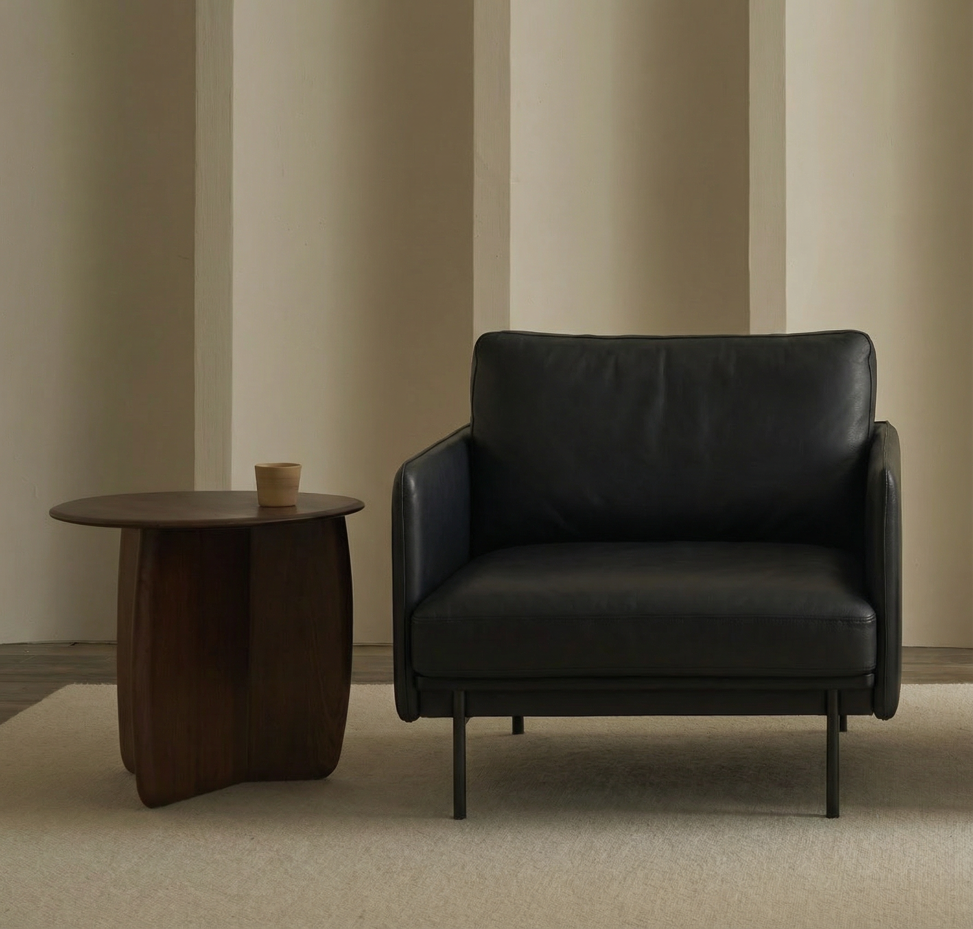 A modern Pensive Lounge Chair | Leather by Wooden Stories in black sits on a light rug beside a round wooden side table with a cup. Beige vertical wall panels in the background create a minimalistic and cozy atmosphere.