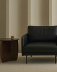 A modern Pensive Lounge Chair | Leather by Wooden Stories in black sits on a light rug beside a round wooden side table with a cup. Beige vertical wall panels in the background create a minimalistic and cozy atmosphere.