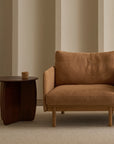 A modern, minimalist living room featuring the Pensive Lounge Chair | Fabric by Wooden Stories with Danish-inspired lines, paired with a dark wood side table holding a cup, set against a neutral wall and light rug.