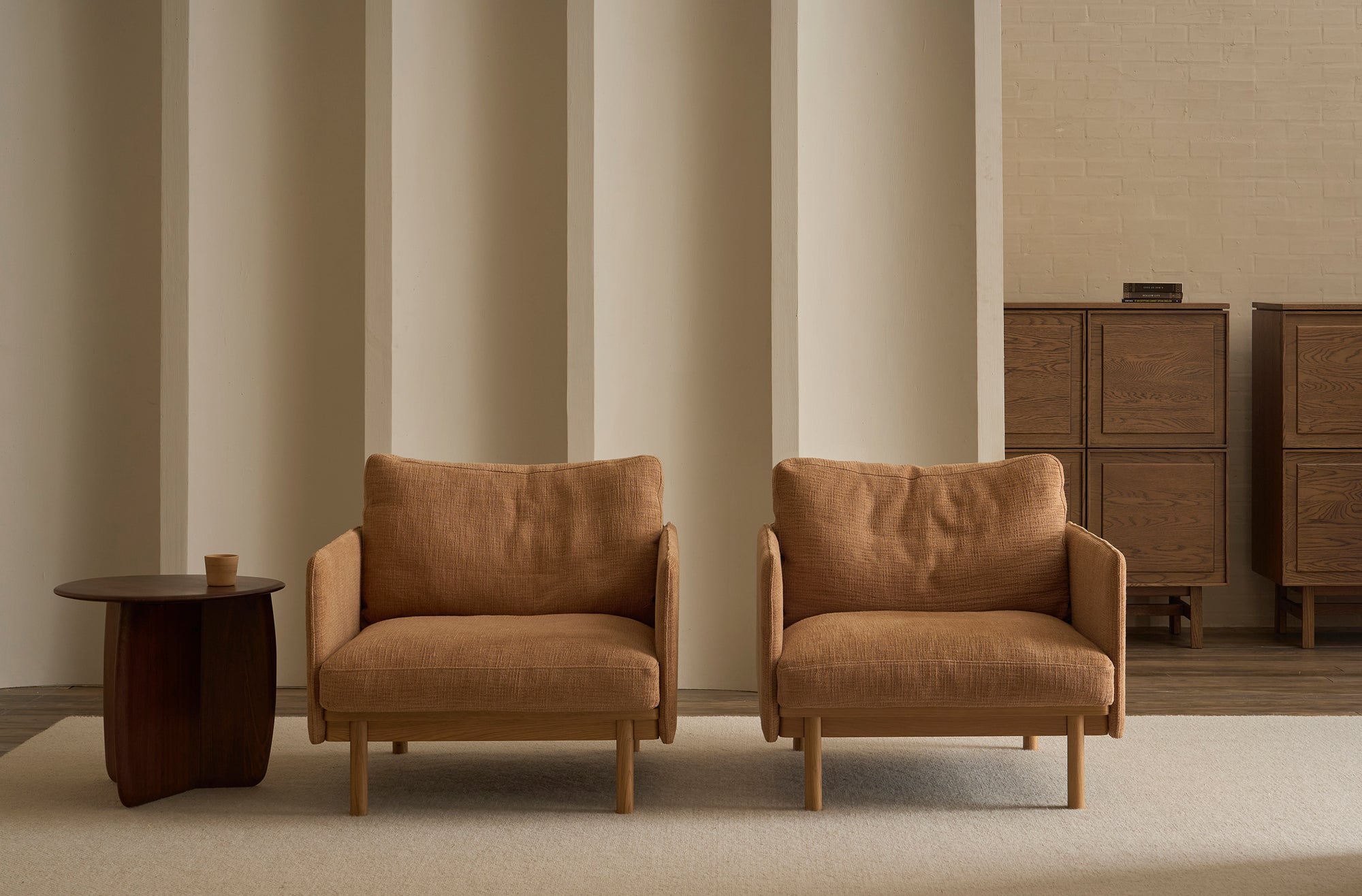 Two tan Pensive Lounge Chairs by Wooden Stories sit side by side on a light rug next to a small dark wood side table, with two tall wooden cabinets against a beige vertically paneled wall in the background.