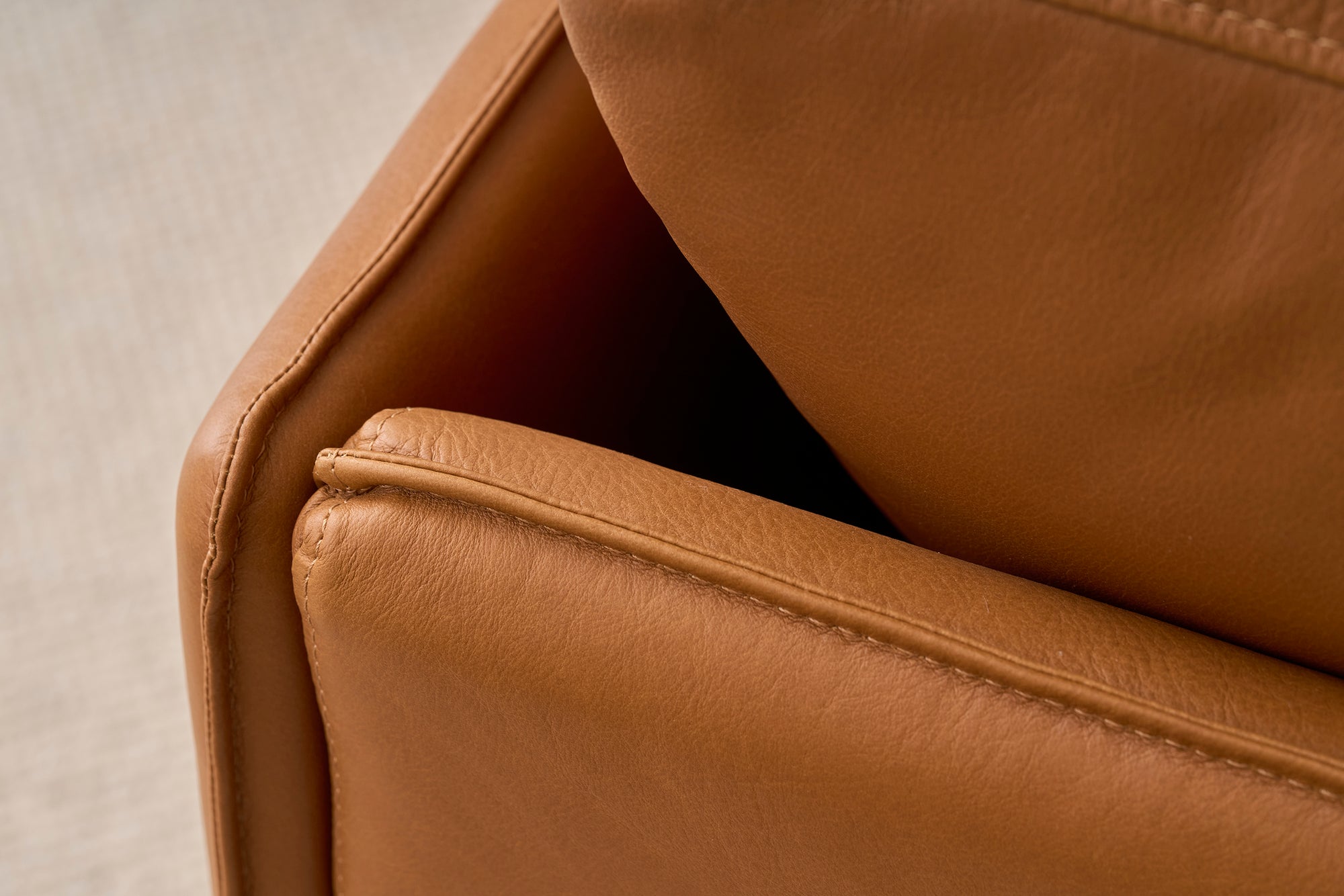 Close-up of a corner of the Pensive Sofa | Leather by Wooden Stories, highlighting its smooth, textured brown leather and detailed stitching. The background features a softly blurred, light-colored carpet.