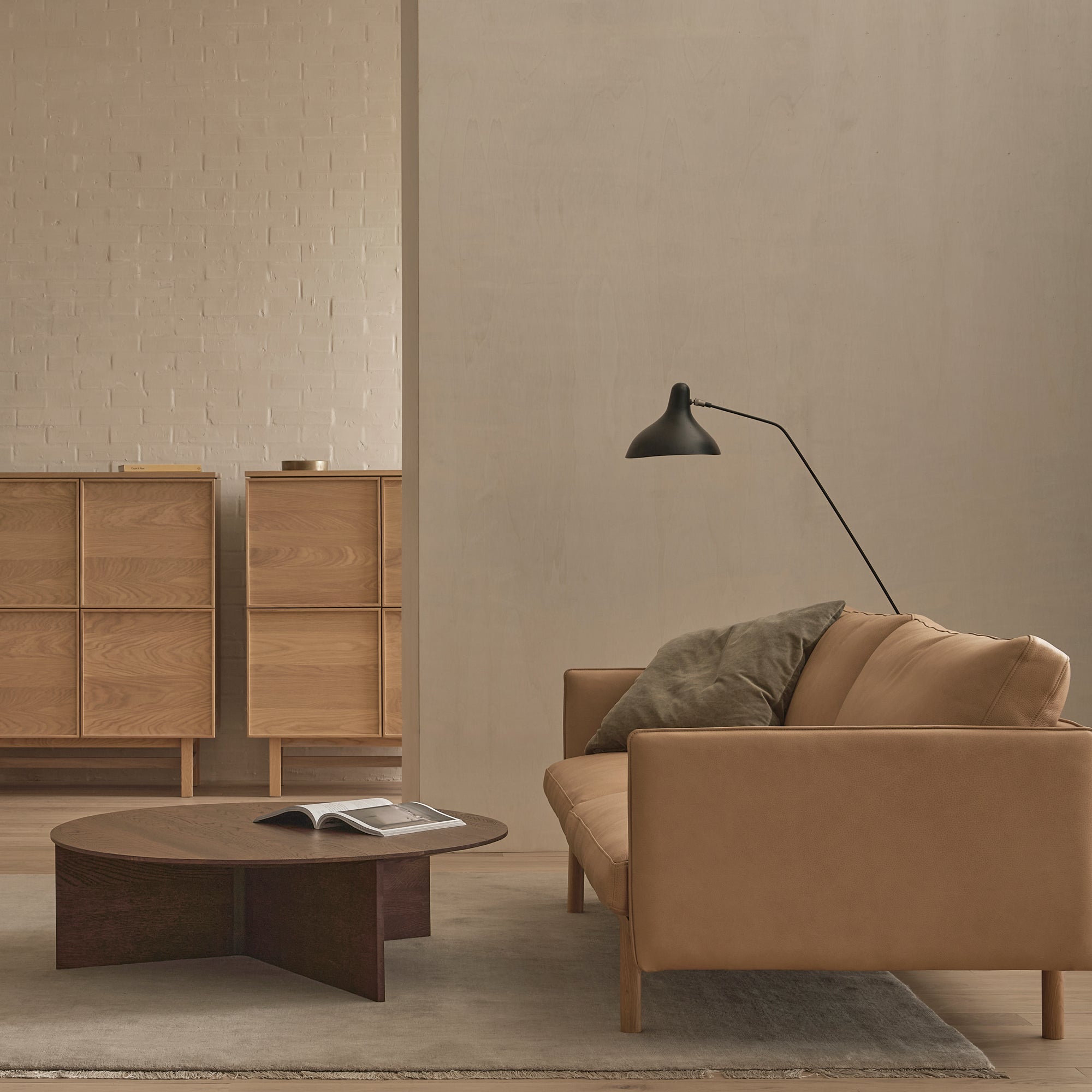 A minimalist living room features the Wooden Stories Pensive Sofa in tan leather, paired with a dark wood coffee table, an open book, two wooden storage cabinets, a gray cushion, and a black floor lamp set against neutral walls.
