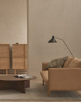 A minimalist living room features the Wooden Stories Pensive Sofa in tan leather, paired with a dark wood coffee table, an open book, two wooden storage cabinets, a gray cushion, and a black floor lamp set against neutral walls.