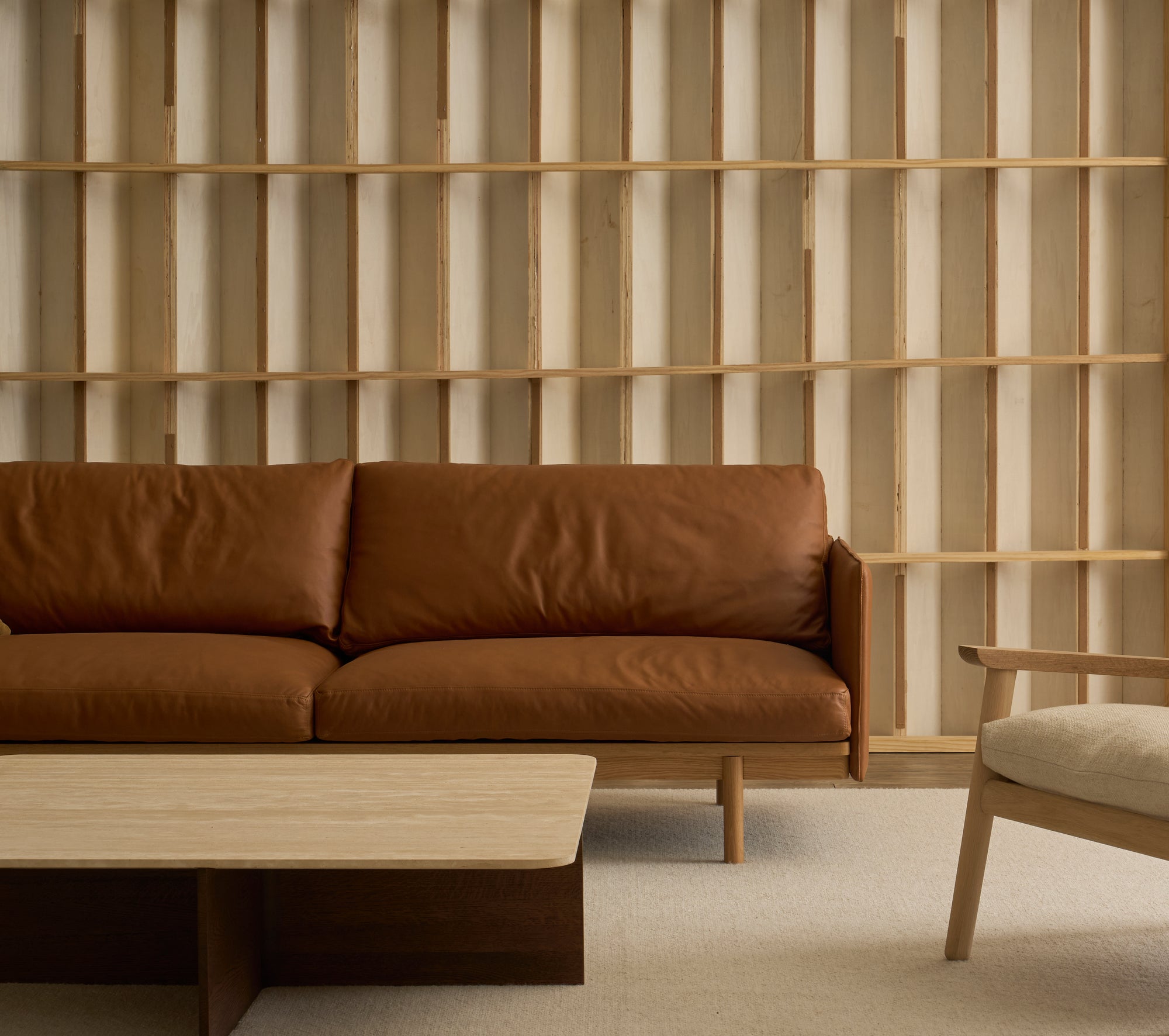 Minimalist living room featuring the Wooden Stories Pensive Sofa in brown leather, a light wood coffee table, beige armchair, and a wooden slat wall. Neutral colors and clean lines create an inviting atmosphere.
