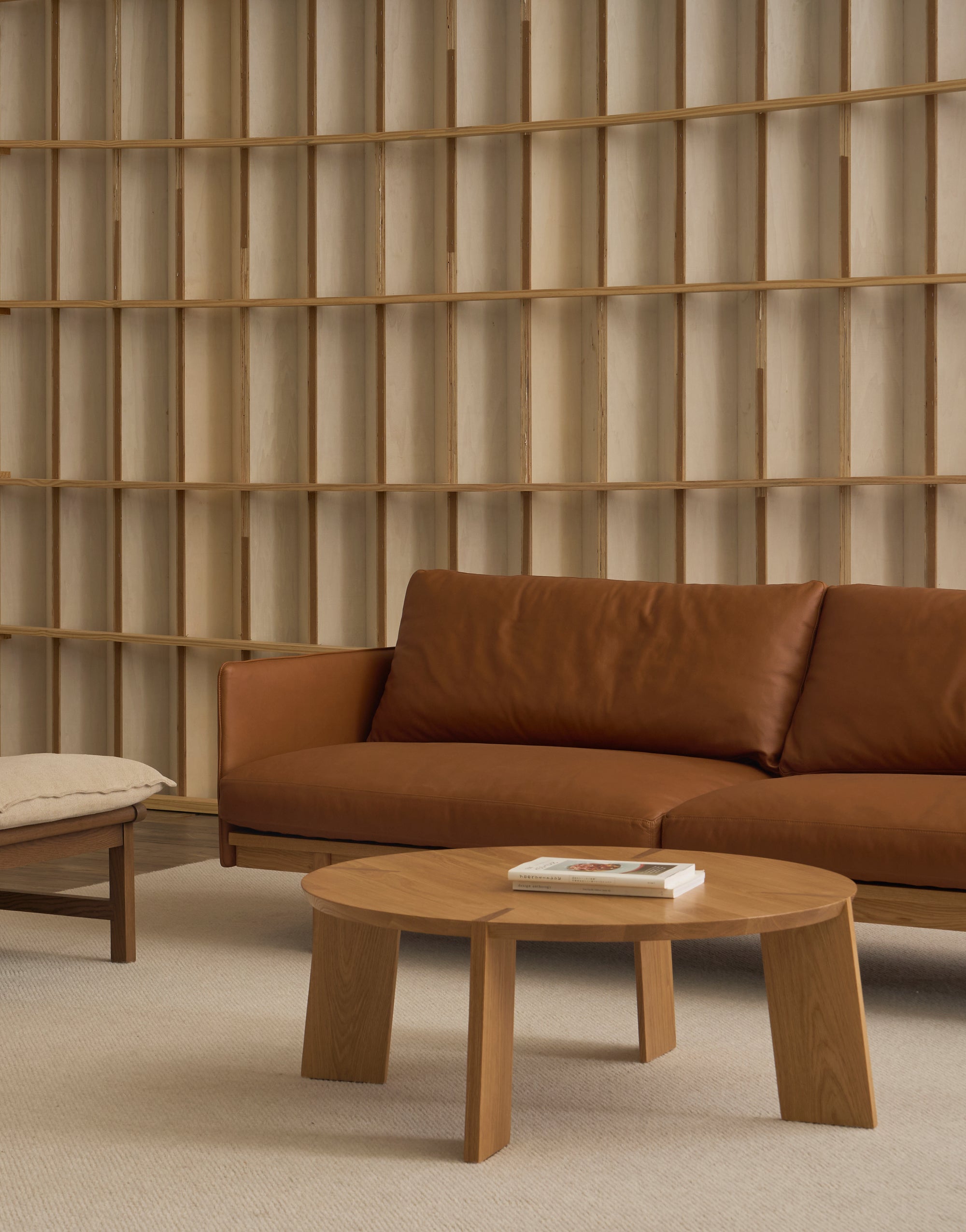 A minimalist living room with the Wooden Stories Pensive Sofa in brown leather, a light wood round coffee table with books, a beige cushioned bench, and a decorative wooden grid wall in the background.