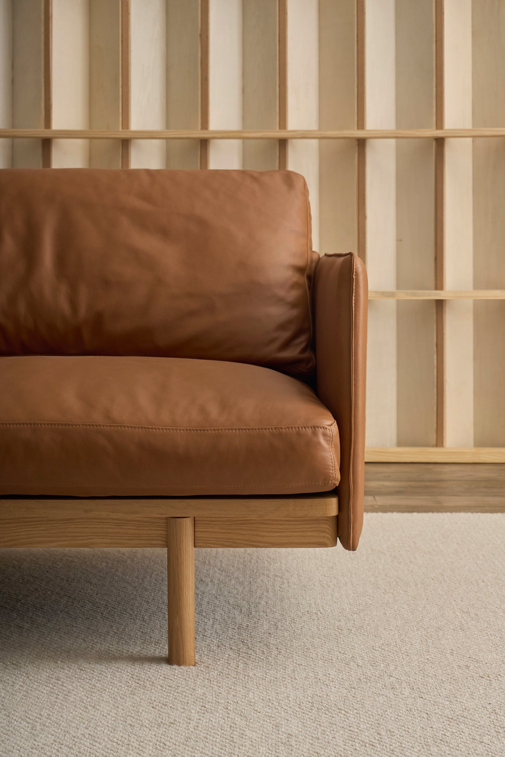 The Pensive Sofa | Leather by Wooden Stories features modern brown leather upholstery with a wooden frame and legs, set against a light wood slat wall and neutral textured carpet.