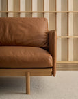 The Pensive Sofa | Leather by Wooden Stories features modern brown leather upholstery with a wooden frame and legs, set against a light wood slat wall and neutral textured carpet.