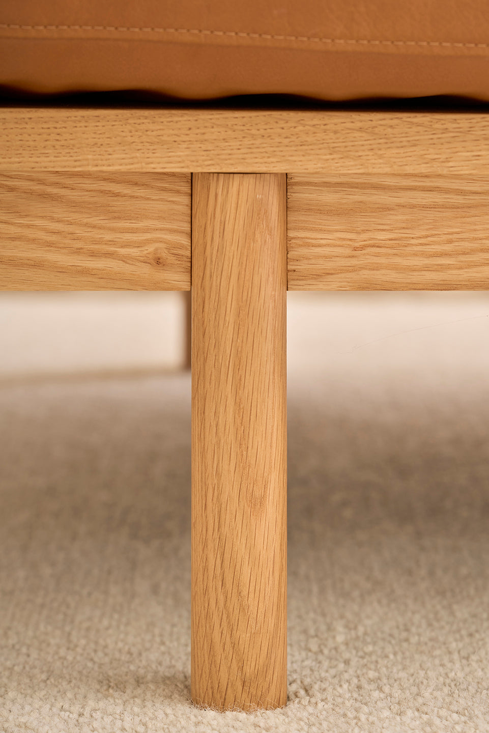Close-up of a wooden leg attached to the frame of the Pensive Sofa | Leather by Wooden Stories, shown on a light textured carpet with a tan leather seat above.