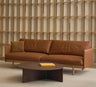 A modern living room featuring Wooden Stories' Pensive Sofa | Leather in brown, a matching pillow, a round dark wood coffee table with a book, and Danish collection wooden shelving in the background.