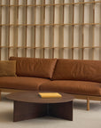 A modern living room featuring Wooden Stories' Pensive Sofa | Leather in brown, a matching pillow, a round dark wood coffee table with a book, and Danish collection wooden shelving in the background.