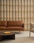 A modern living room featuring the Wooden Stories Pensive Sofa in brown leather, a light beige armchair, a round dark wood coffee table with a closed yellow book, and a decorative wooden slat wall in the background.