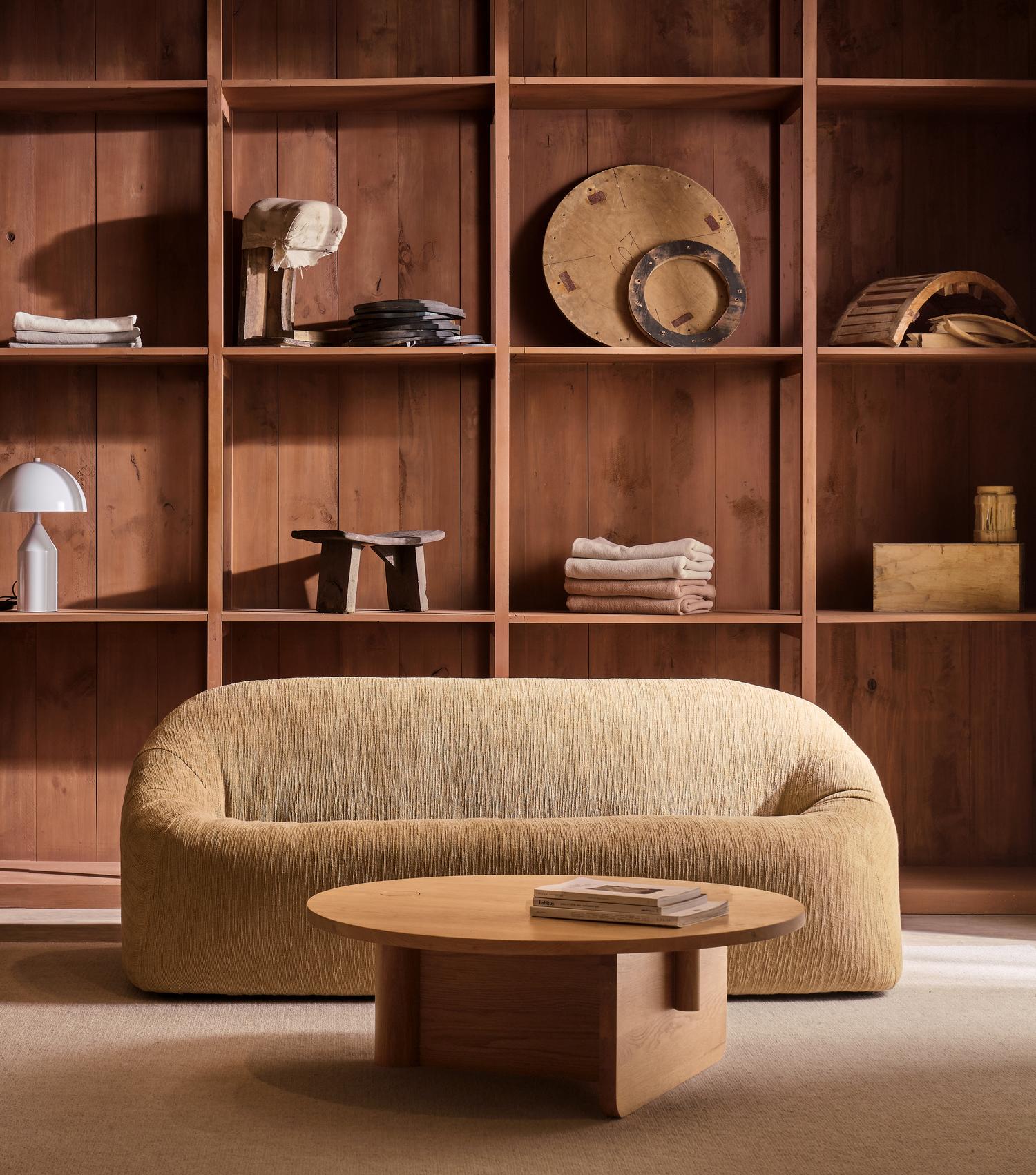 A cozy beige sofa faces a wooden bookshelf filled with towels, books, and decor. In front of the sofa, the Pivot Coffee Table | Round - Wood from Wooden Stories rests on a light carpet, topped with books.