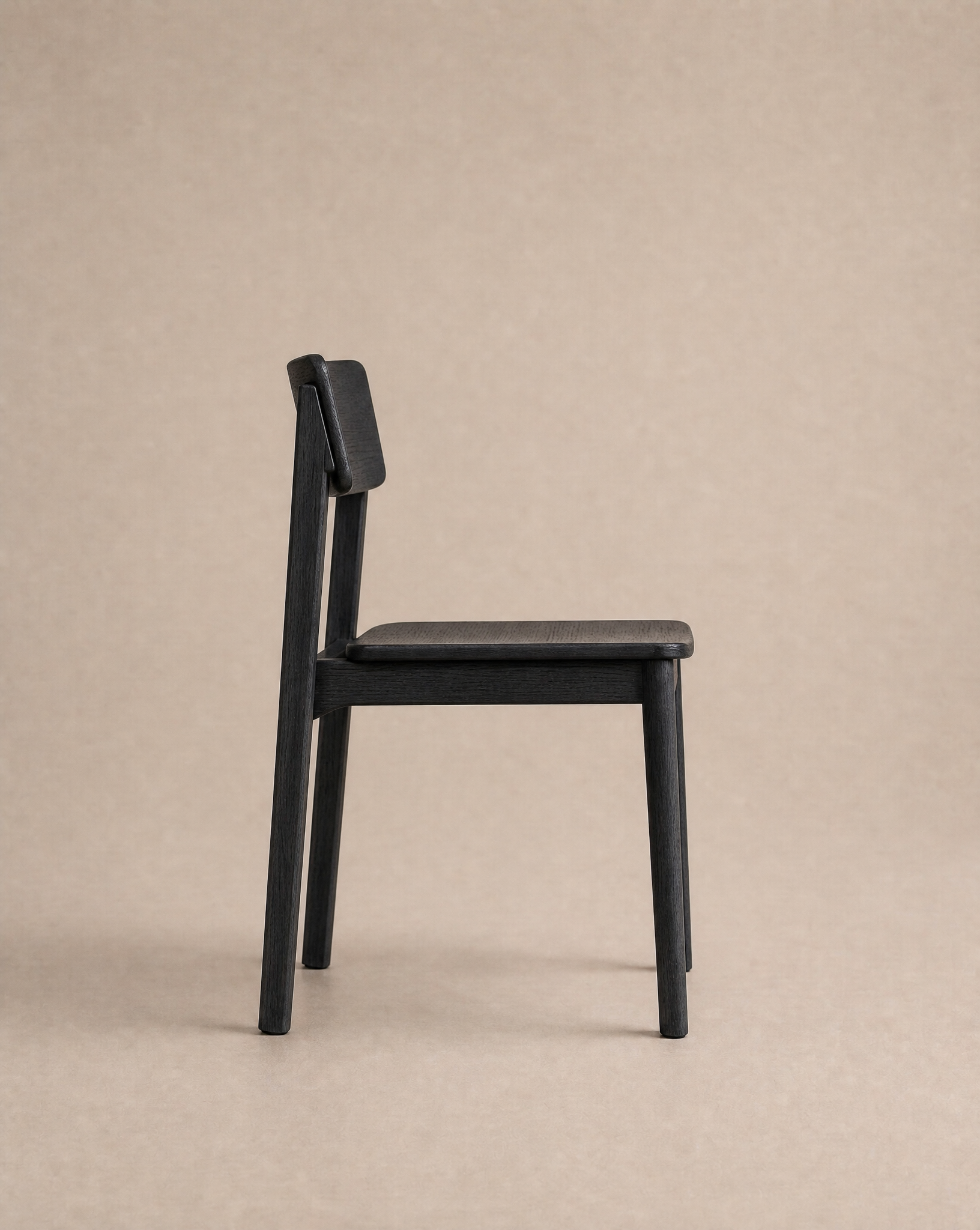 The Poise Dining Chair | Wood by Wooden Stories is shown in profile against a plain beige background. This minimalist black solid oak chair features a simple, modern design with straight legs and an angled backrest, making it contract grade and suitable for both residential and commercial spaces.