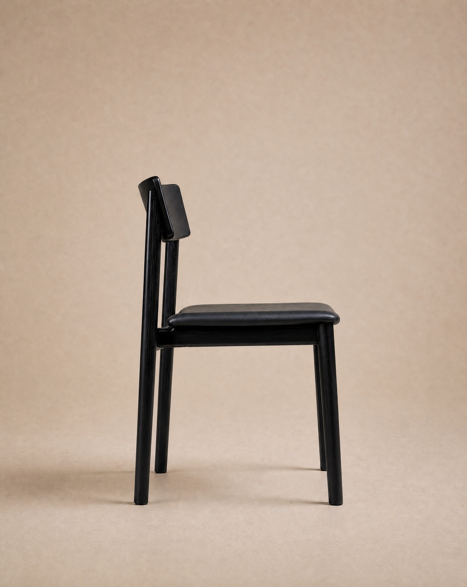 Here’s a revised version of your sentence using the provided product data: The Poise Dining Chair | Leather by Wooden Stories, featuring a modern black wooden frame with a cushioned leather seat and angled backrest, is shown in profile against a plain beige background.