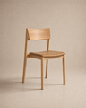 The Poise Dining Chair | Leather by Wooden Stories features a minimalist solid oak frame with a curved backrest and a flat leather seat, presented against a neutral beige background.