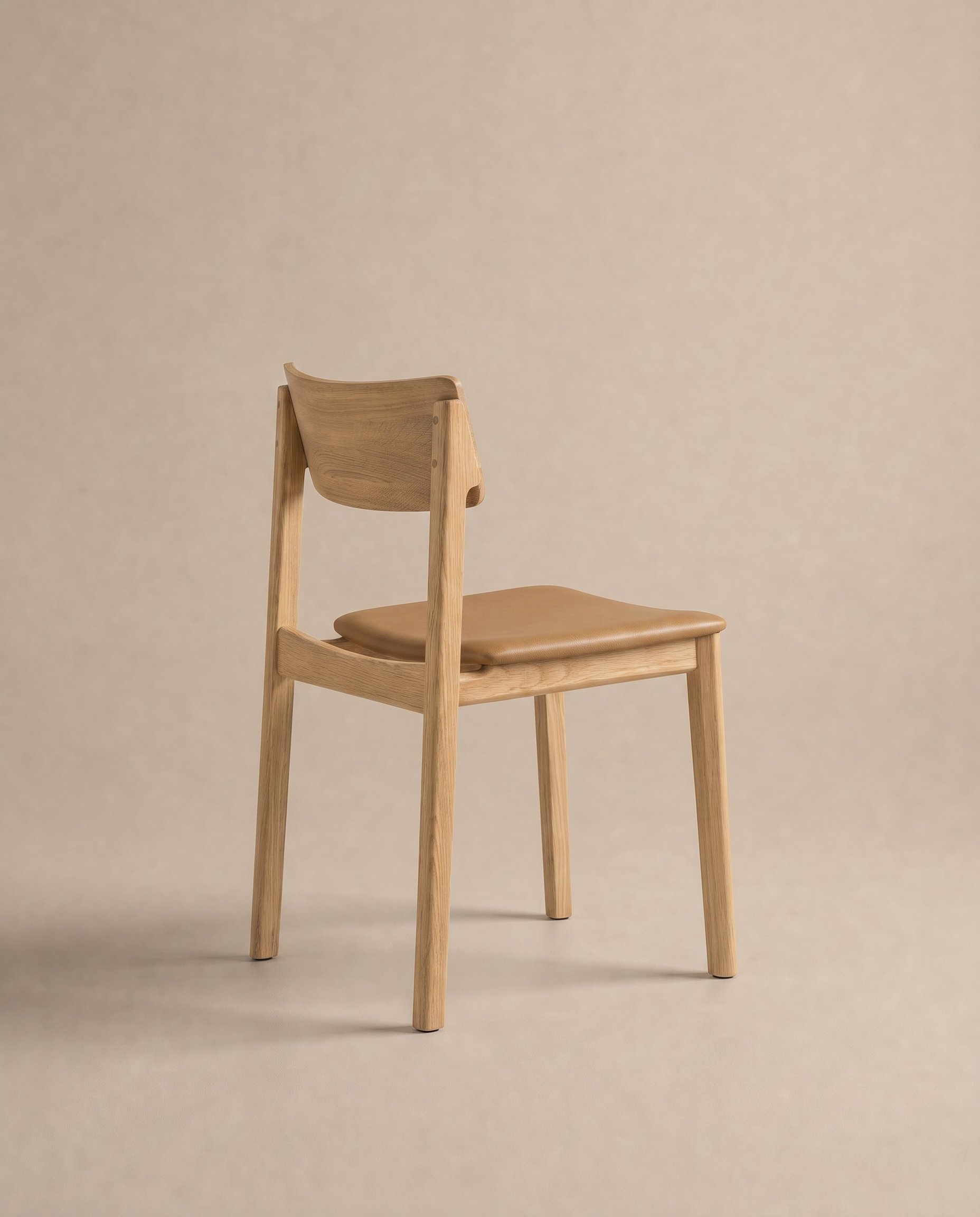 A simple wooden dining chair from Wooden Stories’ Danish Collection, the Poise Dining Chair | Leather features a solid oak frame with light brown padded leather seat and backrest. It is photographed against a plain beige background, angled to reveal its back and side.