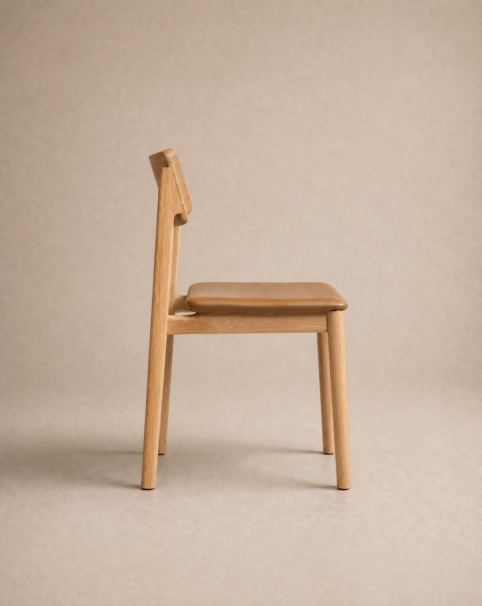 The Poise Dining Chair | Leather by Wooden Stories, featuring a solid oak frame and a tan leather cushioned seat with a simple, modern design, is shown in profile against a neutral beige background.