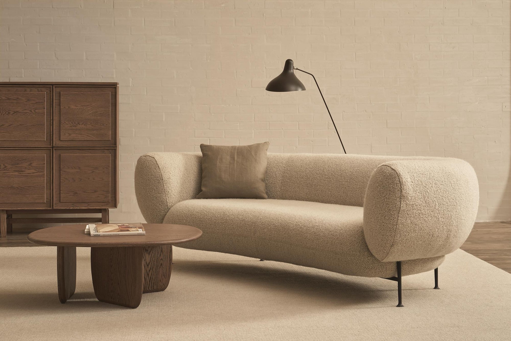 A modern, minimalist living room featuring a cream-colored boucle sofa with a matching throw pillow, the Wooden Stories Eden Coffee Table in Espresso Oak with books, a wooden cabinet, and a black floor lamp against a light brick wall.