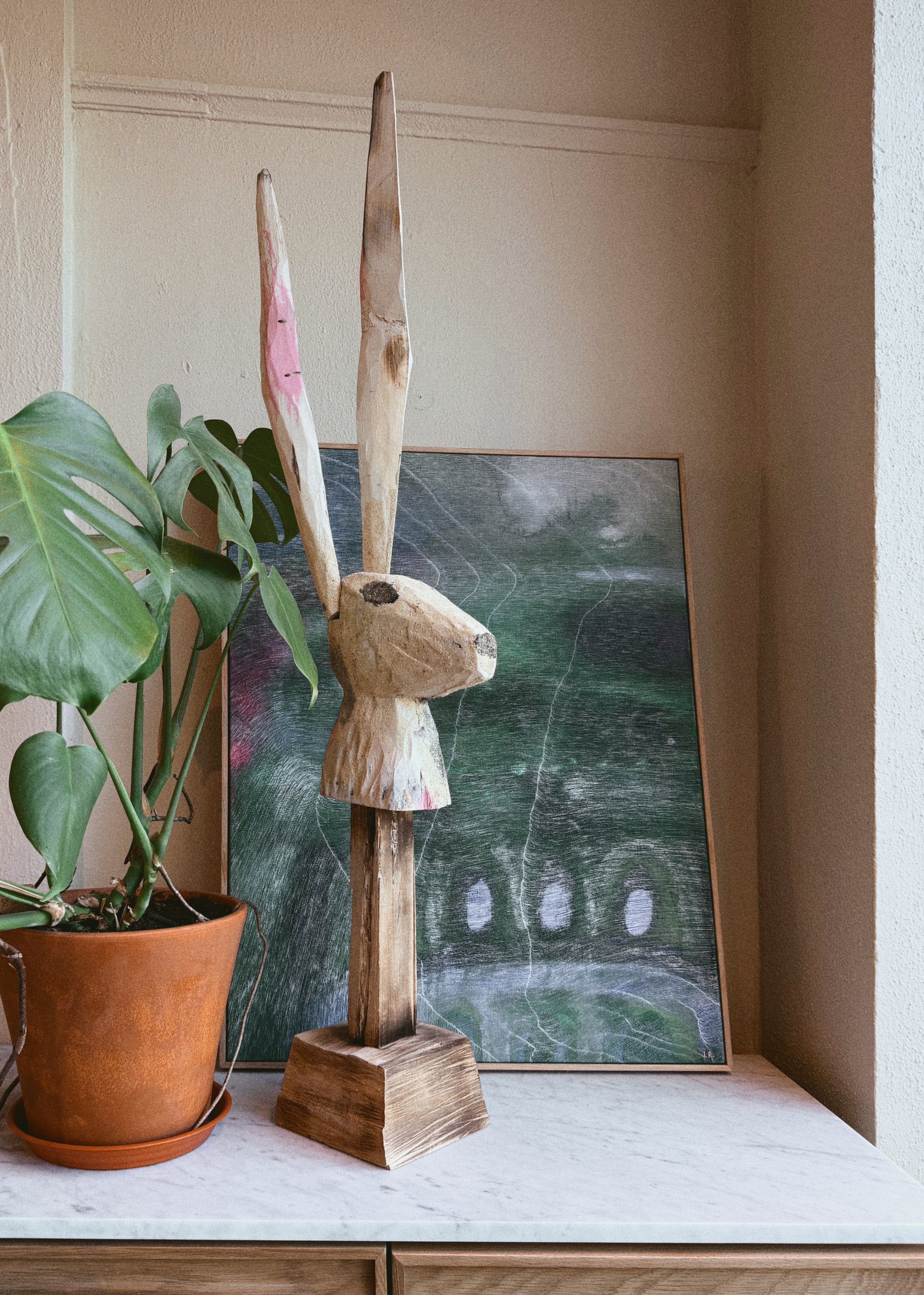 The "Rabbit by Robert Brady" wood sculpture by ROBERT BRADY stands on a white surface beside a potted monstera, with a green abstract framed artwork in the background, echoing the artist’s signature figurative abstraction style.