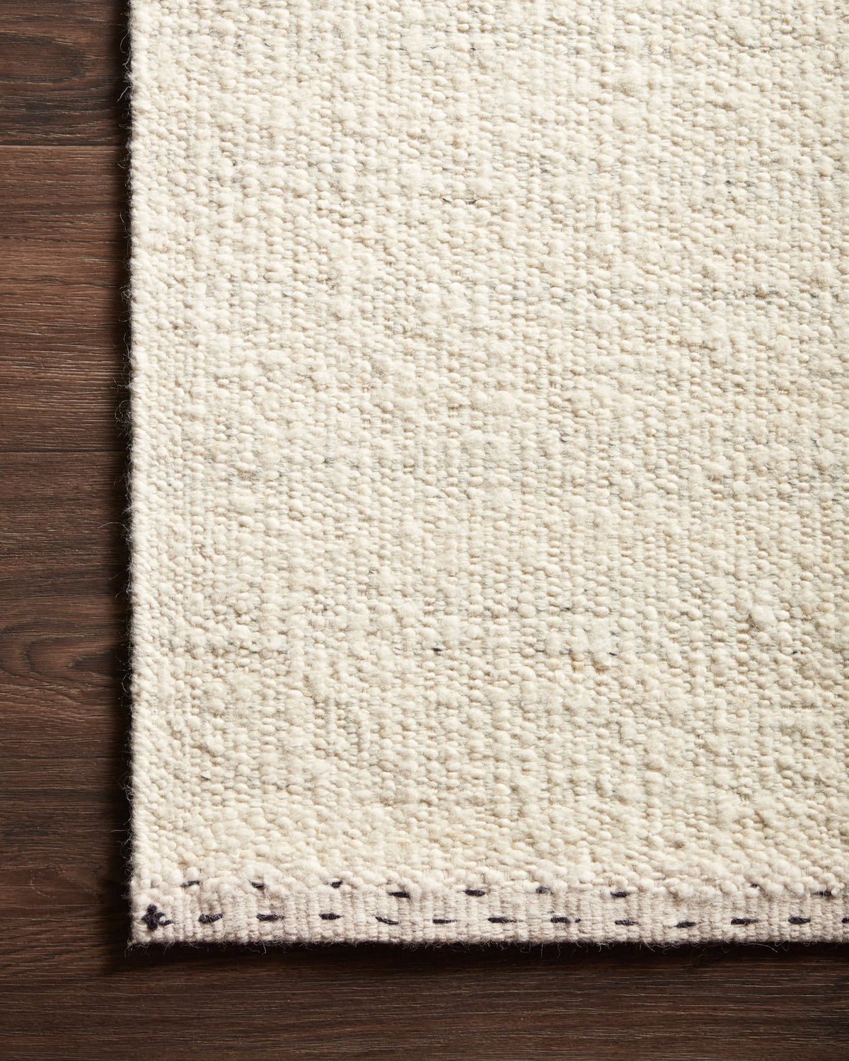 A close-up of the corner of the Sloane Rug - Mist by Loloi, showcasing its textured cream-colored hand-woven design with small dark stitches along the edge, placed on a dark wooden floor.