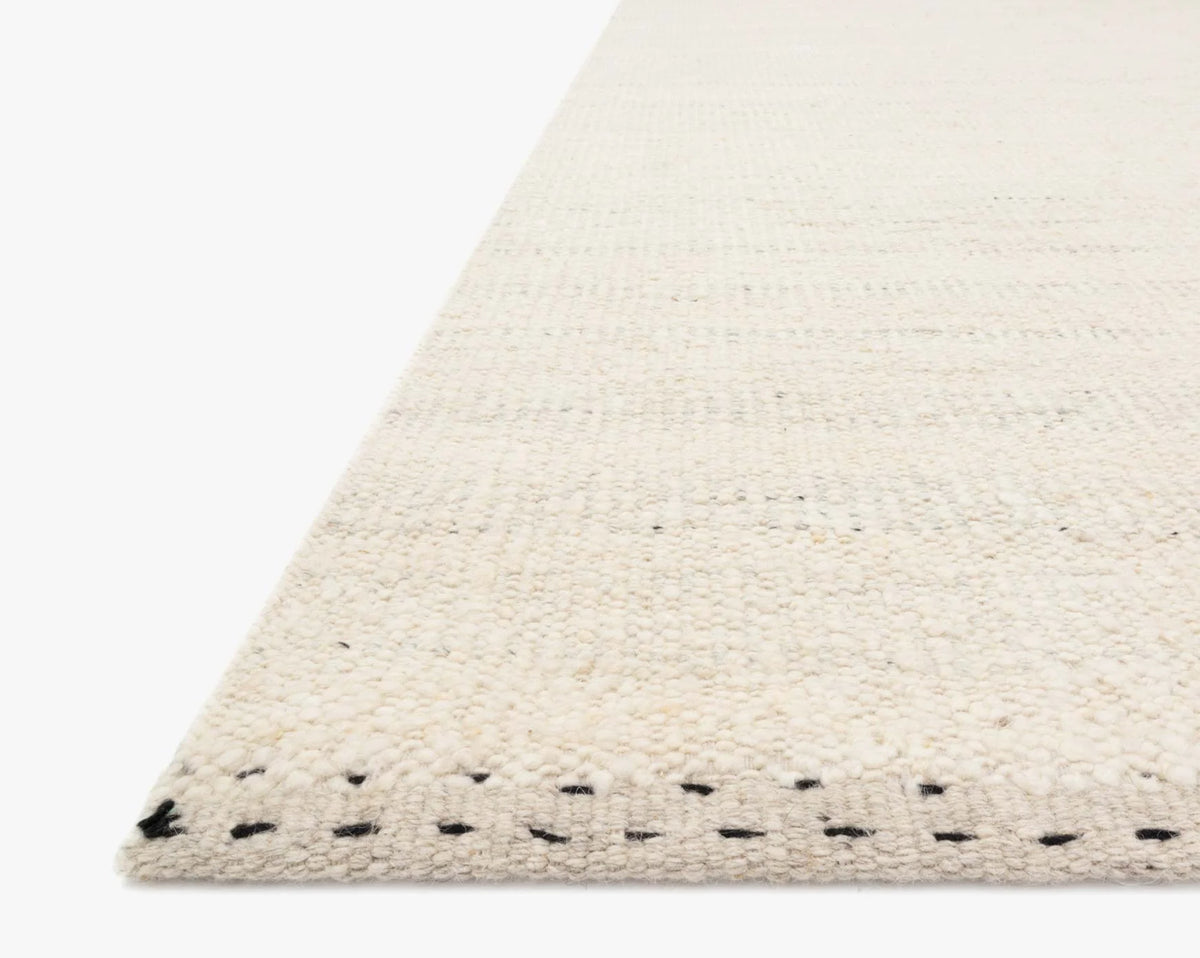 Close-up side view of the Sloane Rug - Mist by Loloi, a hand-woven, off-white rug featuring a textured finish and subtle black stitch accents along the edge. This contemporary bohemian piece from the Sloane Collection is displayed on a white background.