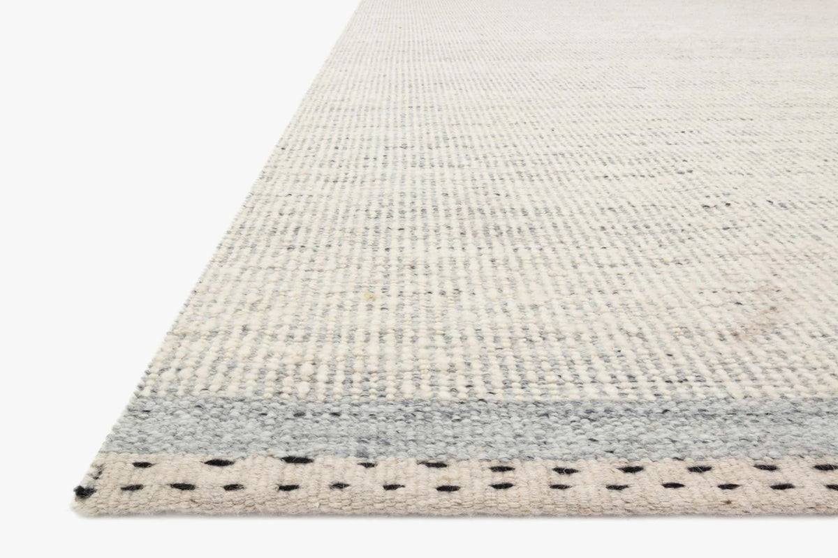 A close-up of the Loloi Sloane Rug - Sky showcases its light, hand-woven texture with subtle gray stripes and a dotted border, set against a white background. This contemporary bohemian rug seamlessly combines pattern and texture for a stylish finish.