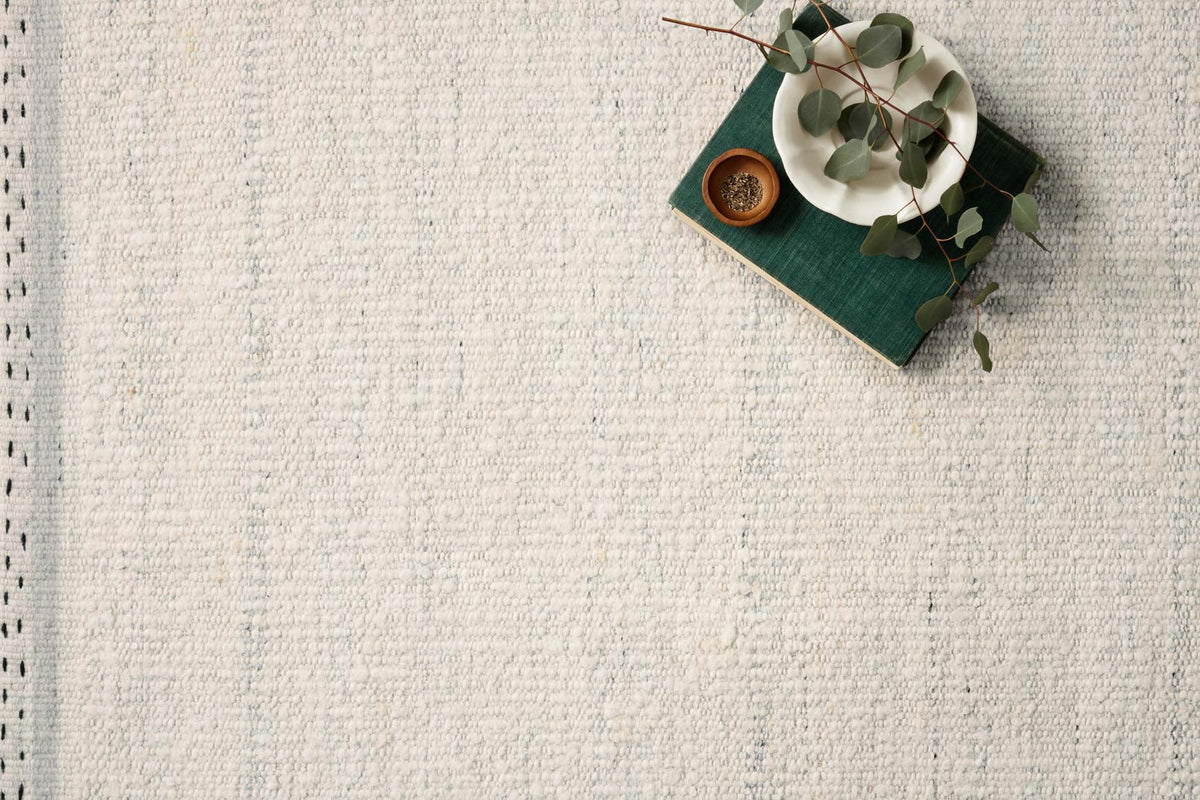 A hand-woven white textured Sloane Rug - Sky by Loloi is shown with a green book, a white plate with leaves, and a small brown dish arranged in the top right corner.