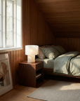 A cozy bedroom with wood-paneled walls, a large bed with green bedding, a wooden nightstand featuring the Snegl Scallop Table Lamp from RUSSO BETAK STUDIO, a beige rug, and abstract artwork resting on the floor by a bright window.