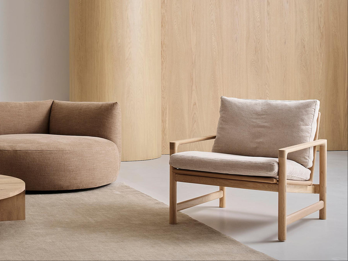 Minimalist living room with a beige curved sofa, matching cushioned armchair, the Troubadour Lounge Chair by Wooden Stories in fabric, a light wood coffee table displaying a small centerpiece, and light wood-paneled walls, creating a calm, neutral-toned space.