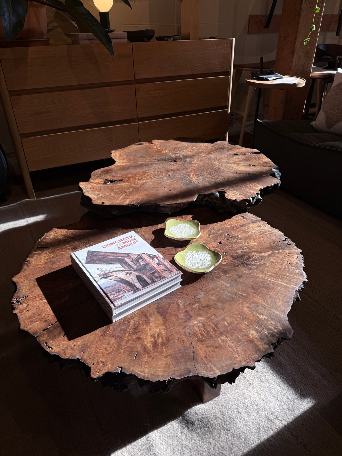 The Bolinas Nesting Coffee Table Set - Claro Walnut by SOBU features two handcrafted coffee tables with natural edges, made in Oakland. In a sunlit living room furnished with wood and neutral decor, stacked books and two small decorative plates adorn their surfaces.