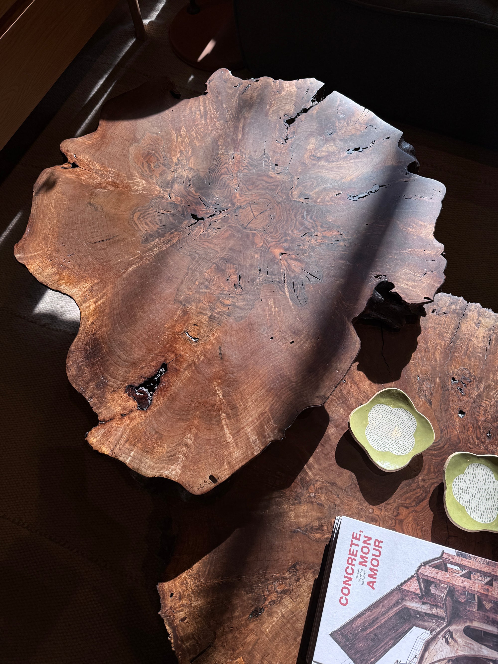 A spacious, rustic Bolinas Nesting Coffee Table Set in Claro Walnut by SOBU, featuring a natural live edge and handcrafted in Oakland, glows in the sunlight. Nearby, two small green dishes and the book titled CONCRETE, MEMOIR, AMOUR rest on another wooden surface.