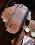 A spacious, rustic Bolinas Nesting Coffee Table Set in Claro Walnut by SOBU, featuring a natural live edge and handcrafted in Oakland, glows in the sunlight. Nearby, two small green dishes and the book titled CONCRETE, MEMOIR, AMOUR rest on another wooden surface.