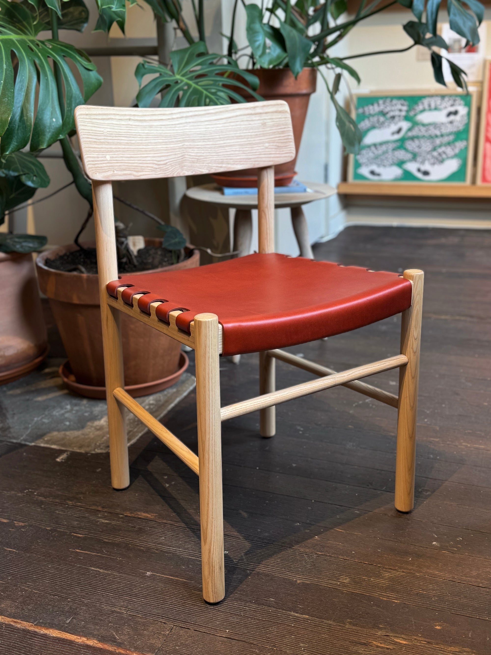 A SOBU Cesco Dining Chair in solid white ash with a heritage red leather seat sits on a dark wooden floor, surrounded by potted plants and colorful framed artwork in the background.