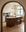A modern kitchen with a marble-topped island, SOBU’s Cesco Counter Stools in walnut featuring top grain leather seats, gold fixtures, a large window, and a decorative fireplace—all viewed through an American black walnut archway. The space boasts minimalist decor in neutral tones and elegant lighting.