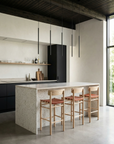 A modern minimalist kitchen with a large stone island, three SOBU Cesco Counter Stools in White Ash topped with top grain leather seats, black cabinets, open shelves with dishes, slim pendant lights, and sunlight streaming through a large window.